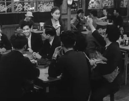 An old woman in the bar selling cigarettes approaches a table full of students, who rebuff her