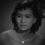 A black-and-white photo of a Japanese girl in a low-cut gown, looked anxiously off to one side