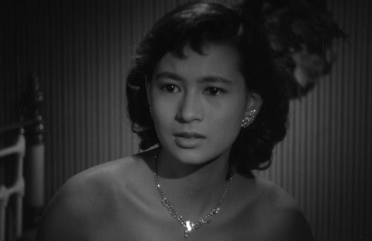 A black-and-white photo of a Japanese girl in a low-cut gown, looked anxiously off to one side