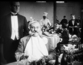 Photo of a white-haired man in a crowded restaurant, clearly wealthy, being waited on while other diners are visible in the background