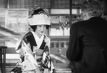 In an interior scene, a Japanese woman in a traditional wedding costume kneels before a man wearing a suit in the foreground, back to camera