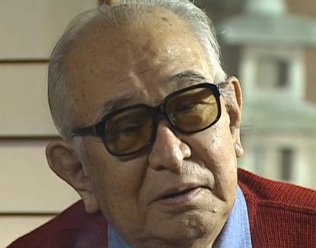 Photo of an elderly Japanese man, Akira Kurosawa, in a red sweater and classes, who appears to be engaging in a conversation with someone off-camera