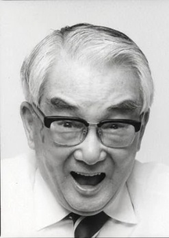 An elderly Japanese man with glasses in a shirt and tie, looking as if he's about to speak