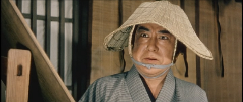 A middle-aged man with a disfigured face, Jirō, wearing a sedge hat that partially conceals his fact, looks on the procession with rage and horror