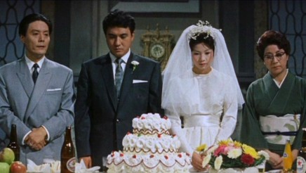 Four modern Japanese people at a wedding, with the bride in a Western bridal gown and the groom in the center, flanked by an older man, in a suit, and woman, in a kimono; there is a wedding cake and flowers on the table in front of them