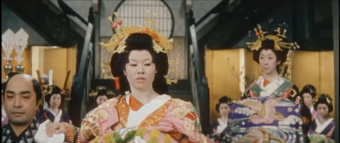 Two Japanese women in the dress of ancient courtesans both face the camera, the one on the right looking at the one in the middle, who ignores her and rests her right hand on the shoulder of a male bodyguard to her right