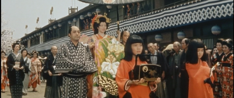 Two little girls dressed in red lead the procession in 18th Century Yoshiwara, the older carrying a box, the younger a scroll; a courtesan flanked by guards follows, and a middle-aged man and woman follow after her, as bystanders watch