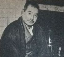 Medium close photo of a middle-aged Japanese man in traditional garb, sitting in a kitchen with bottles of wine