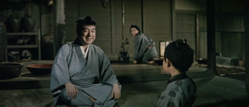 A domestic scene in ancient Japan: a middle-age blind man, smiling, talks to a little boy facing away from us, while a woman watches in the background