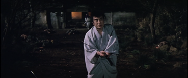 In ancient Japan, a middle-aged man standing outdoors at night brandishes a sword, while a woman whose face is covered watches from the background