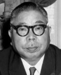 Portrait photo of an elderly Japanese man, with grey hair, large black glasses and wearing a business suit, looking unsmiling into the camera