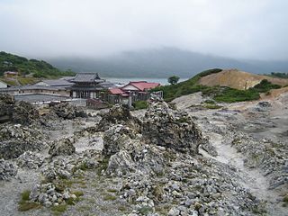 Photo of part of Mount Osore in Japan, with rock formations in the foreground and a temple and various other structures in the background