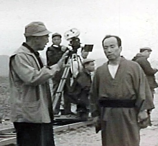 A tall, elderly Japanese man in a coat and hat talks to a middle-aged man wearing a traditional kimono, while cameras and film personnel are visible in the background