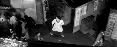 A woman, seen from overhead, runs through a street of shops, looking backward in fear