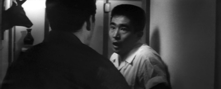 A young Japanese man in casual clothes looks with terror at the older man, Tarumi, who is approaching him, back to camera 