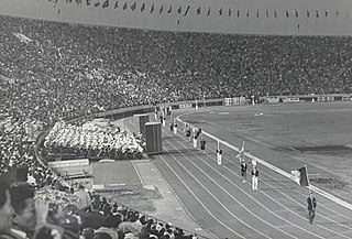 Photo of a stadium with stands full of people, and a handful of people in the far distance marching on the playing field, in the final ceremony of the 1964 Tokyo Olympics