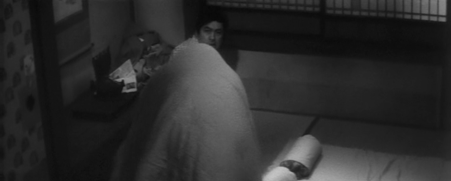 A woman covered up in a blanket and thus not visible, jokingly looms over a man, Inukai, sitting on the floor, who seems to be not at all amused