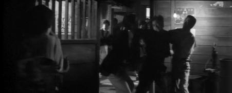 A woman in an apron, Yae, watches from behind a partition as a young yakuza, Machida, punches another gang member in the stomach with the help of a friend