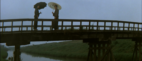 On a bridge in the near distance, two Japanese men carrying umbrellas wearing traditional dress are conversing 