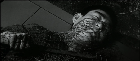 Black-and-white shot of a young Japanese man lying dead on the ground, half his face obscured by netting