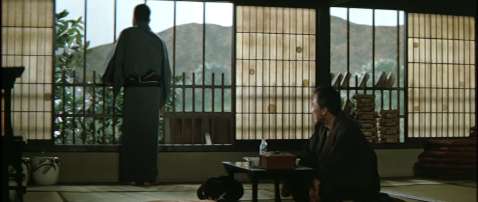 Two Japanese men dressed in traditional kimono, with the younger man, back to camera, looking out the window at mountains in the distance, while the older man sits on the floor