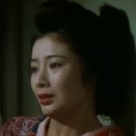 Head and shoulders shot of a young Japanese woman in traditional dress leaning against a wall, looking distraught