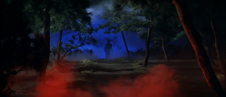 A color photo in which a man in silhouette walks away in the distance, as the night sky behind him is an intense shade of blue and a red mist rises from the ground 