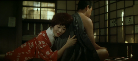 A Japanese woman in traditional clothing, sitting on the floor, embracing a Japanese man, also sitting, so that his kimono is pulled back, exposing his shoulder