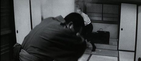 An older Japanese man in the background of the photo flings dirt into the face of the younger Japanese man in kimono, who shields his eyes