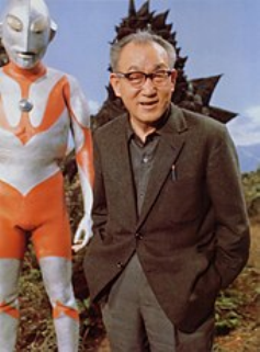 Color photo of a middle-aged Japanese man with glasses and wearing a suit jacket, standing beside a model of the Ultraman character, with the costume of a kaiju character located behind him