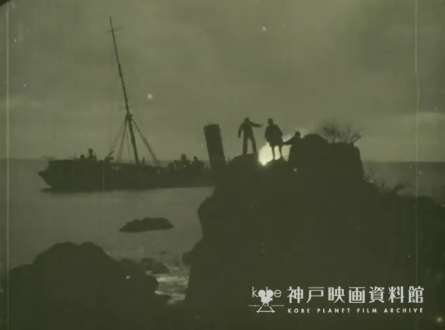 An indistinct image, apparently taken at night, showing a rocky coast with several human figures silhouetted by a bonfire facing a motionless ship on the sea in the far distance.