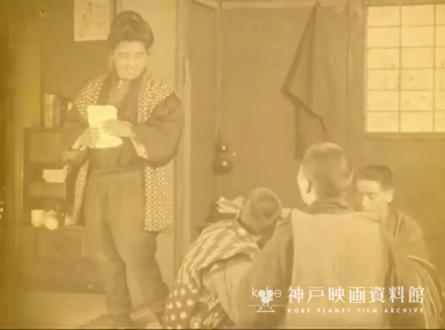 A dimly-seen shot of a young Japanese woman in traditional dress standing in a room holding a package, talking to a group of young boys who are sitting in a circle, watching her