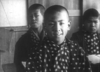 Black-and-white photo of a young Japanese boy with very short hair, sitting in a classroom, with two other boys wearing similar clothes sitting at desks behind him