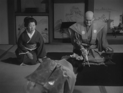 In feudal Japan, in a small room, a tearful young woman prostrates herself before her parents, a middle-aged woman and a slightly older man, who physically dominates both women.