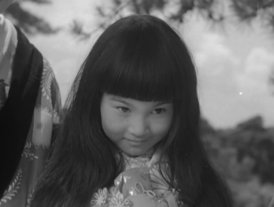 A little girl with long, black hair, in the costume of a child in the feudal era, standing outside in the sun, nods her head and smiles