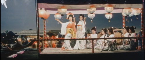 In this color image, a boat is seen floating on a river, while women in traditional Japanese dress dance and fireworks are visible in the background