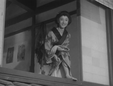 An attractive young woman wearing kimono in a room on the second floor of a house looks down with amusement at something she sees on the street below