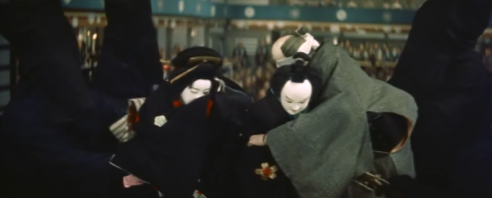 Image of three Japanese Bunraku puppets, during a performance at a puppet theater, from the rear of the stage, with an audience dimly seen in the background