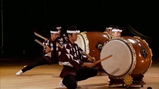 On a stage, two costumed performers play a pair of enormous taiko drums.