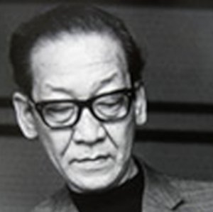 Headshot of an elderly Japanese man in modern clothing wearing black-rimmed eyeglasses, looking down at something in front of him