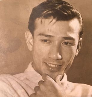 A young Japanese man in modern clothes, smiling, strokes his chin