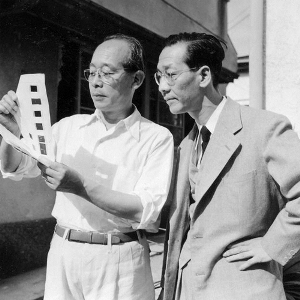 Two Japanese men in modern dress and wearing spectacles, one in jacket and tie and the other in casual clothes, examine what appears to be a strip of film