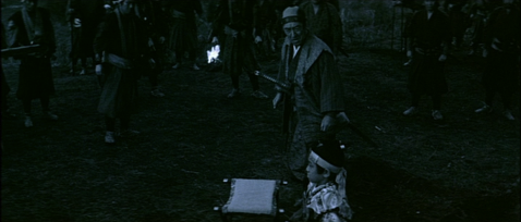 In a rural setting in early morning, a middle-aged man in Japanese costume watches a young boy sitting on a stool near a pine tree, as samurai surround them