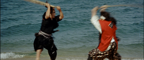 Near the edge of the sea, two Japanese men in feudal costume face each other with their weapons raised, about to attack