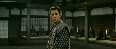 A young Japanese man in feudal costume addresses a group of men in a large hall