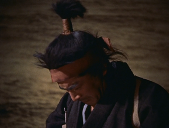 A Japanese man in period costume, with the sea behind him, bows his head in sorrow