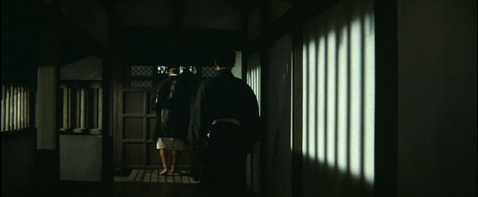 A Japanese man in feudal costume, back to camera, is led down a dark corridor by another man