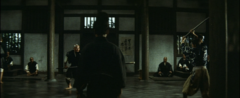 The same young Japanese man as in the previous image, still with his back to the camera, is now in a large hall, watching several men fight with wooden weapons