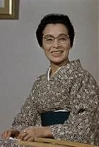 A middle-aged woman in traditional kimono, wearing glasses, smiles at the camera