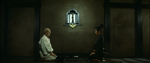 An older Japanese man in a monk's costume and a younger Japanese man in a samurai costume kneel facing each other, a Buddhist statue on the wall between them